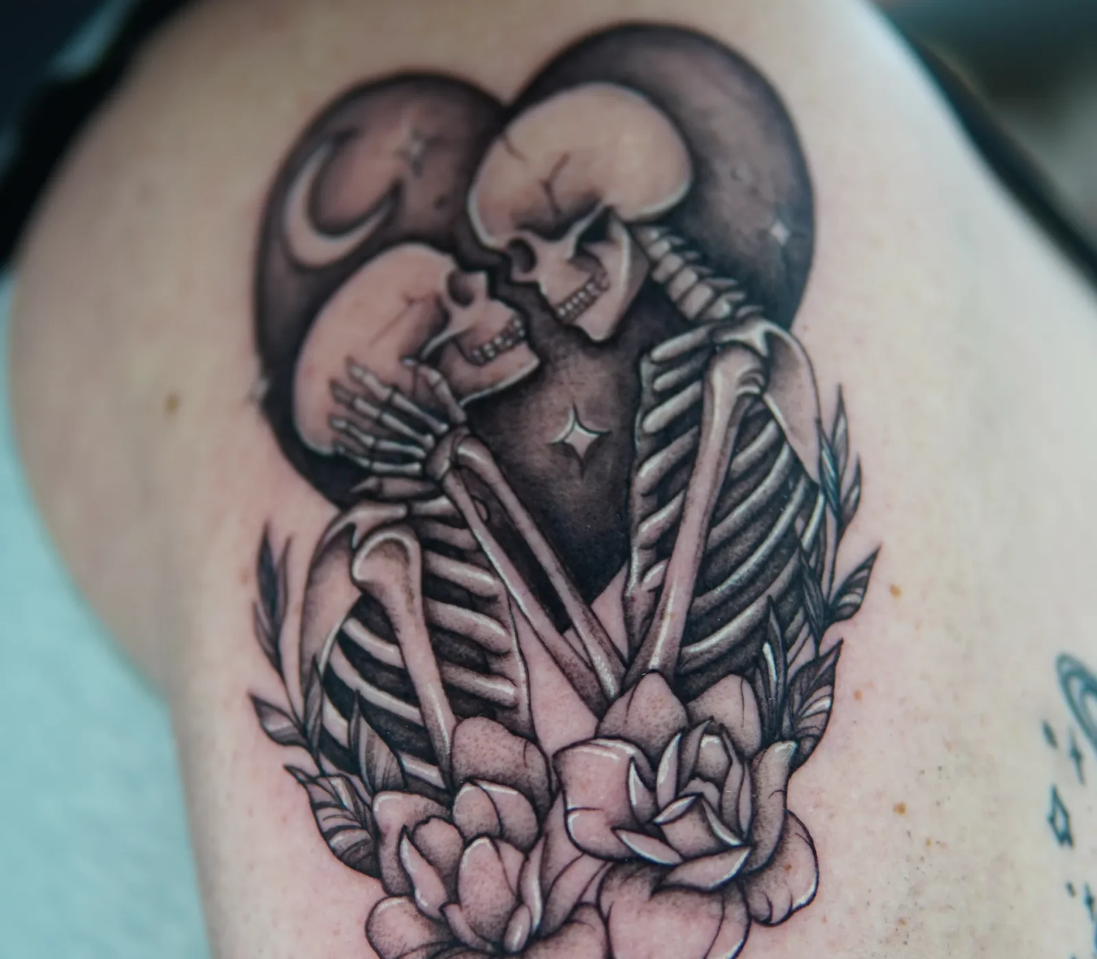Black-and-grey skeleton lovers kissing with flowers tattooed on the upper arm