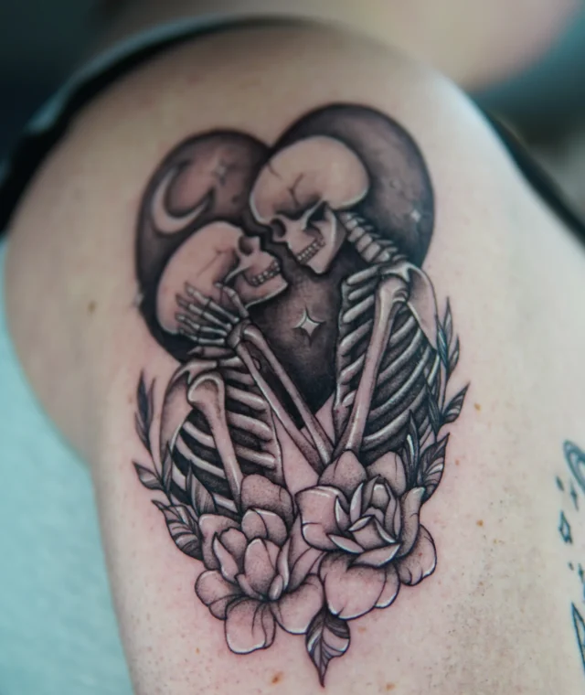 Black-and-grey skeleton lovers kissing with flowers tattooed on the upper arm