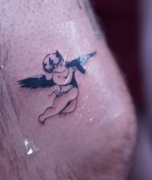 Small winged Cupid reaching toward a bird tattoo near the knee
