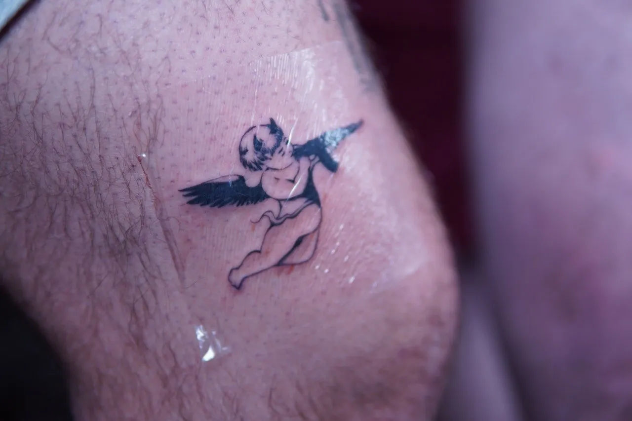 Small winged Cupid reaching toward a bird tattoo near the knee